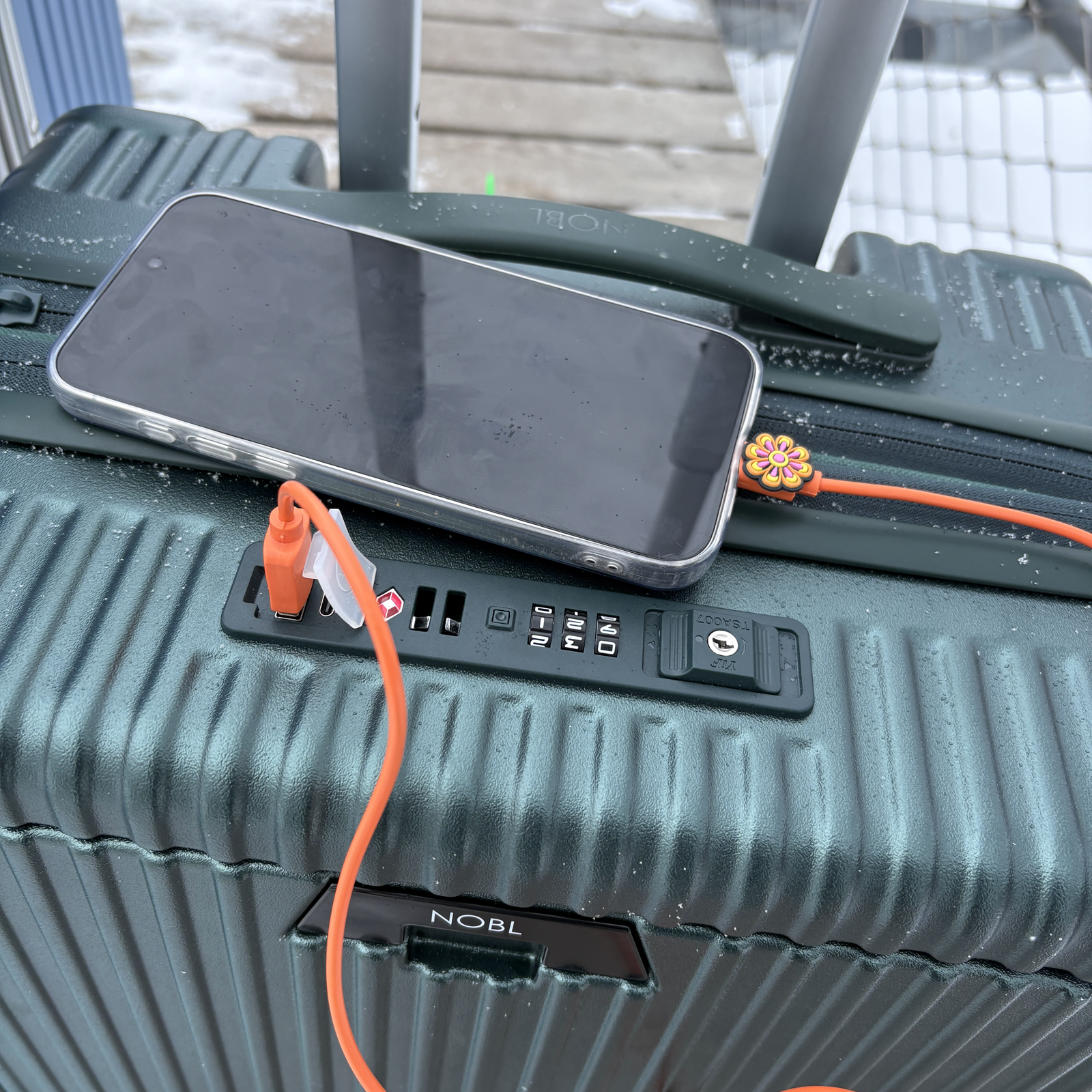Phone charging from the built-in holder on a yellow NOBL carry-on at the airport