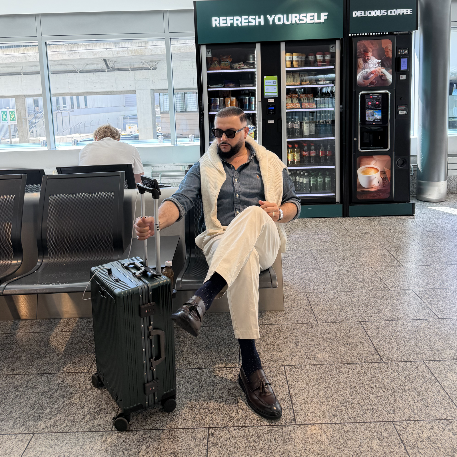 Traveler seated calmly at the airport beside a silver NOBL carry-on and NOBL billboard